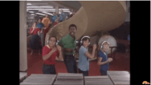 Three children and an adult man dancing in front of a staircase in a library. 6 other adults are dancing on the stairway.