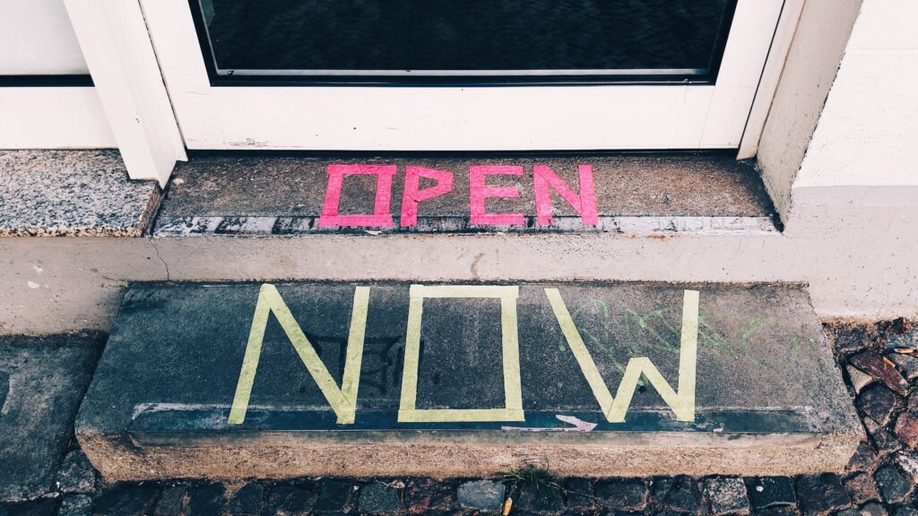 Entryway door with the words "OPEN" and "NOW" written in tape on the two steps leading up to the door, respectively.