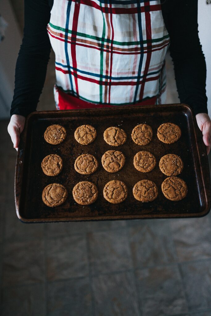 A top down shot of a white adult wearing a plaid apron and holding a baking sheet of freshly baked molasses cookies.