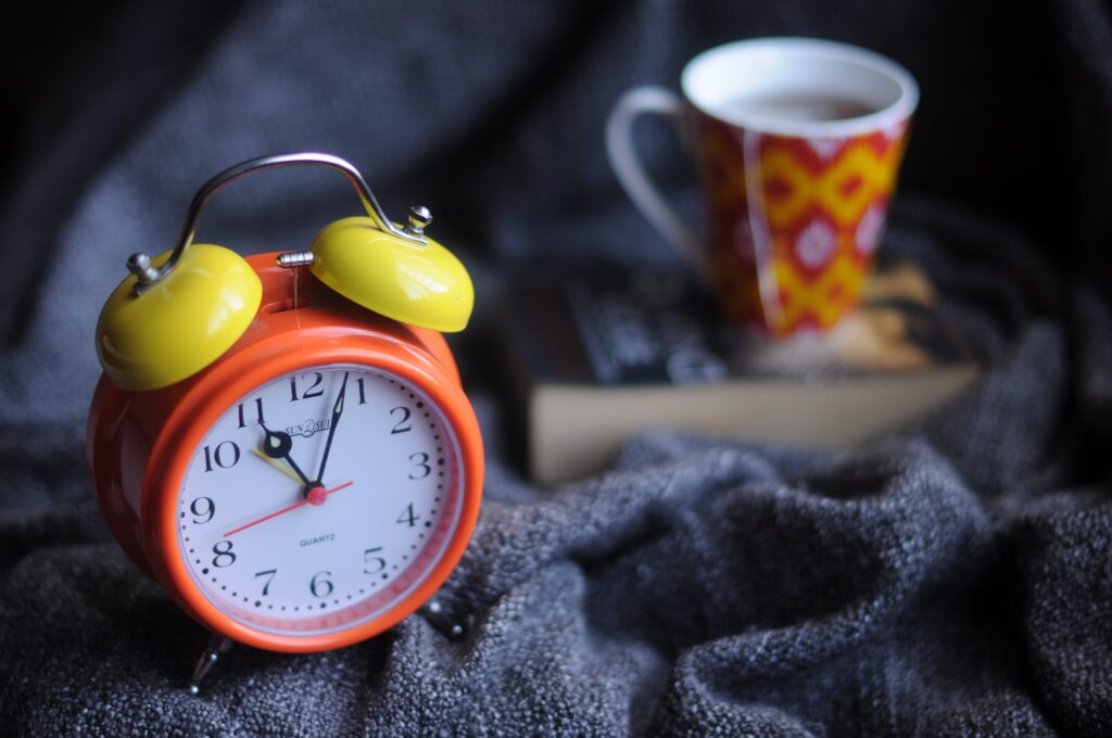 An orange and yellow analog alarm clock sits on a blue cloth in the foreground. A cup of tea sits on top of a paperback book in the background.