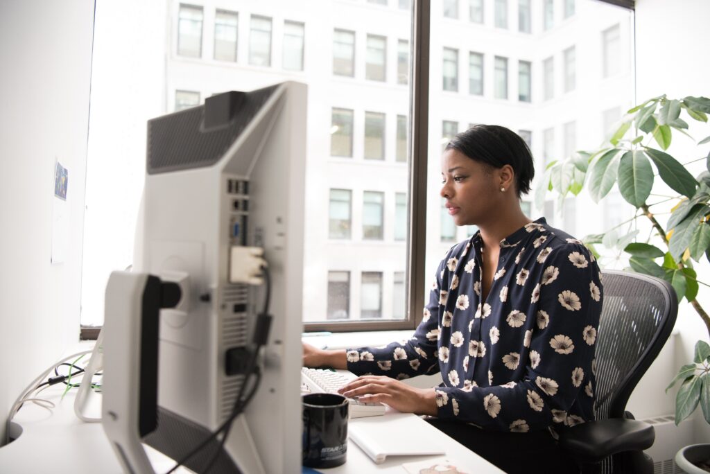 A sitting Black adult woman typing and looking at the two computer monitors in front of her. The white office has tall windows showing the tall buildings behind her.