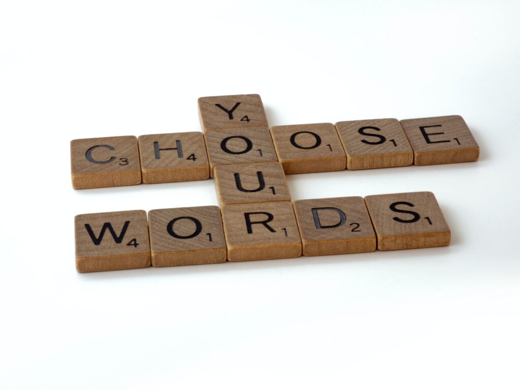 The phase "Choose your words" are spelled out using wooden Scramble letter tiles on a white table. The word "your" is vertically spelled using the "o" and "r" in the horizontal words "choose" and "words", respectively.