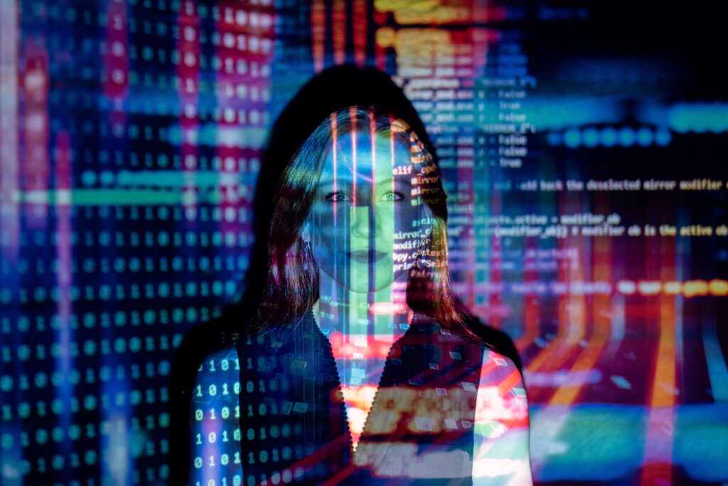 An adult white woman stands in front a wall. Binary code and a programming script are being projected on both the woman and the wall.